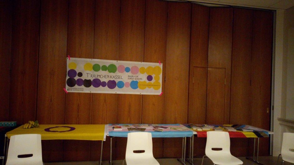 Bild von drei Tischen und Stühlen in einem Veranstaltungsraum. An der holzvertäfelten Wand hängt das bute Logo vom T*räumchen Kassel.