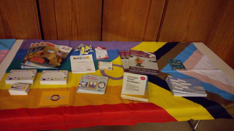Nahaufnahme eines Tisches der von der Regenbogen Pride-Flag bedeckt ist. Darauf liegen diverse Flyer und Postkarten vom T*räumchen Kassel, sowie Info Broschüren rund ums Thema geschlechtliehc Vielfalt.