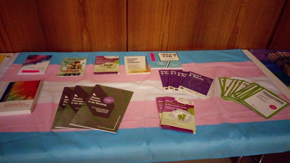 Nahaufnahme eines Tisches der von der pink, weiß, blauen trans* Flagge bedekt ist. Darauf liegen Broschüren für therapeutische Praxis-Tipps rund um das Thema geschlechtliche Vielfalt.