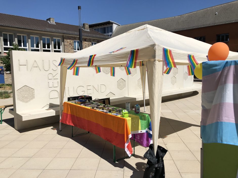 Bild von einem Pavillion-Zelt, das mit Regenbogenflaggen dekoriert ist. Seifenblasen fliegen, eine Trans-Flagge ist an einem Board drapiert, und auf einem Tisch liegen Informationsmaterialien. Hinter dem Zelt ist eine Mauer auf dem der Name des Ortes, Haus der Jugend, zu lesen ist.