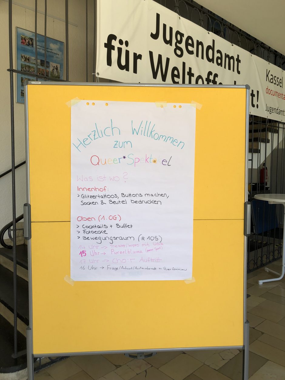 Das Bild zeigt ein händisch gestaltetes Poster auf dem der Programm-Plan und die Orte des Tages angegeben sind. Es ließt: Was ist wo? Innenhof: Glitzertattoos, Bittns machen, Socken und Beutel bedrucken. Oben (1.OG), Cocktails + Buffet, Fotoecke, Bewegungsraum (R 105), 14h Theater- Impro mit Lidia, 15h Purzelblume (queerer Sport), 17h Chor Auftritt, 16h Frage/Antwort/Austausch-Runde zu queeer Feminismus