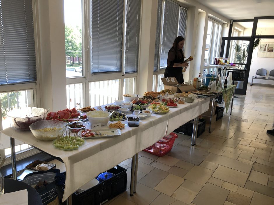 Das Bild zeigt eine Reihe von drei Tischen, die zu einem Buffet und einer Bar zusammen gestellt sind. Auf den Tischen steht viel frisches Essen, wie Obst, Salate, Rohkost und Dips, sowie Cupcakes und Pizzaschnecken. Das Essen ist mit kleinen Schildern versehen die das Essen benennen und verraten ob es vegan ist oder nicht. Hinter der Bar steht eine Person, die Getränke mischt.
